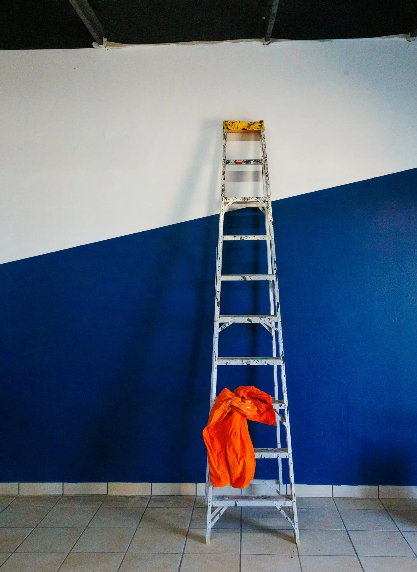Leiter mit orangem Tuch an einer halb blau, halb weiß gestrichenen Wand